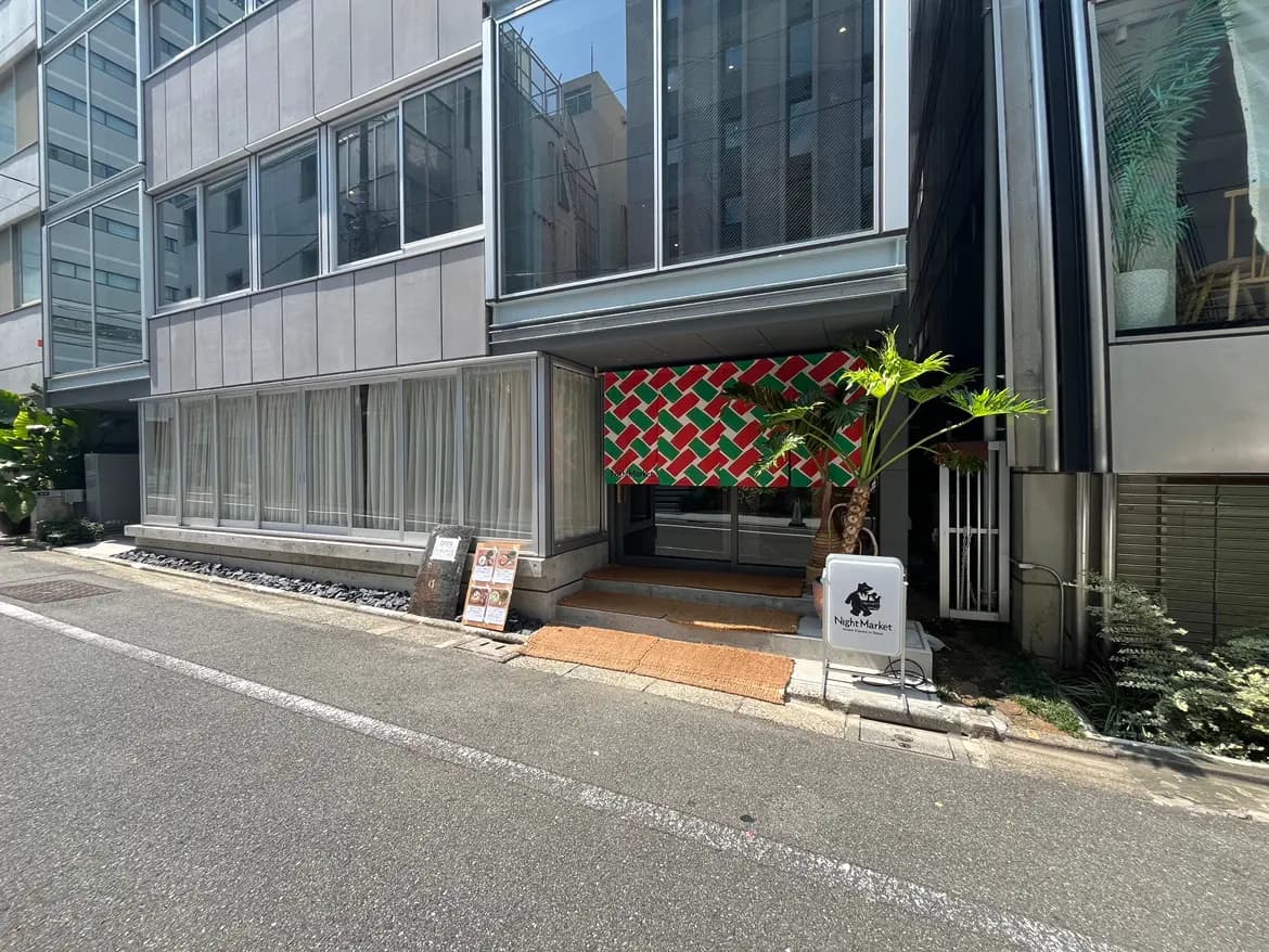 - モダンな建築様式の店舗外観、カラフルなテントと植栽が特徴的なエントランス、周囲に整備された歩道と商業スペース