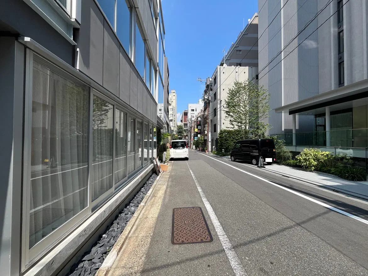 新しい都市型マンションの外観、通り沿いに並ぶ植栽と車両、青空の下で明るい雰囲気の街並み