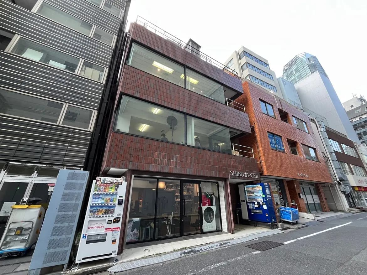- 渋谷エリアに位置するモダンな商業ビルの外観、レンガ調のファサードと大きな窓、通りに面した自動販売機とショップの入り口