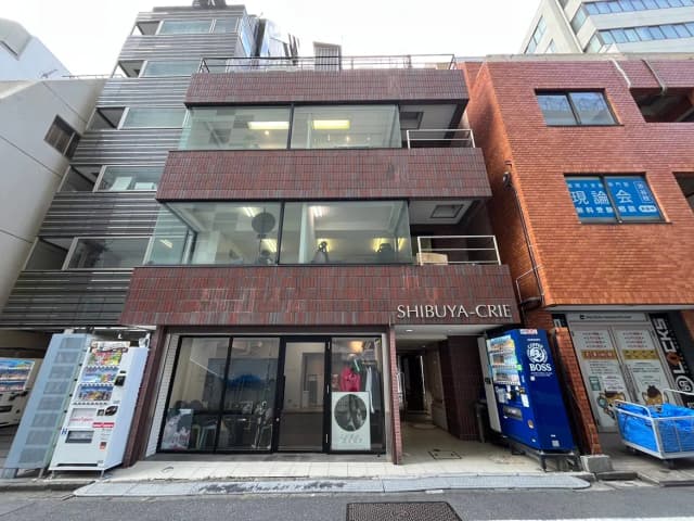 - 渋谷にあるモダンな商業ビル「SHIBUYA-CRIE」、ガラス張りの外観と赤茶色のタイルが特徴的なデザイン、ビル前には自動販売機が並ぶ景観