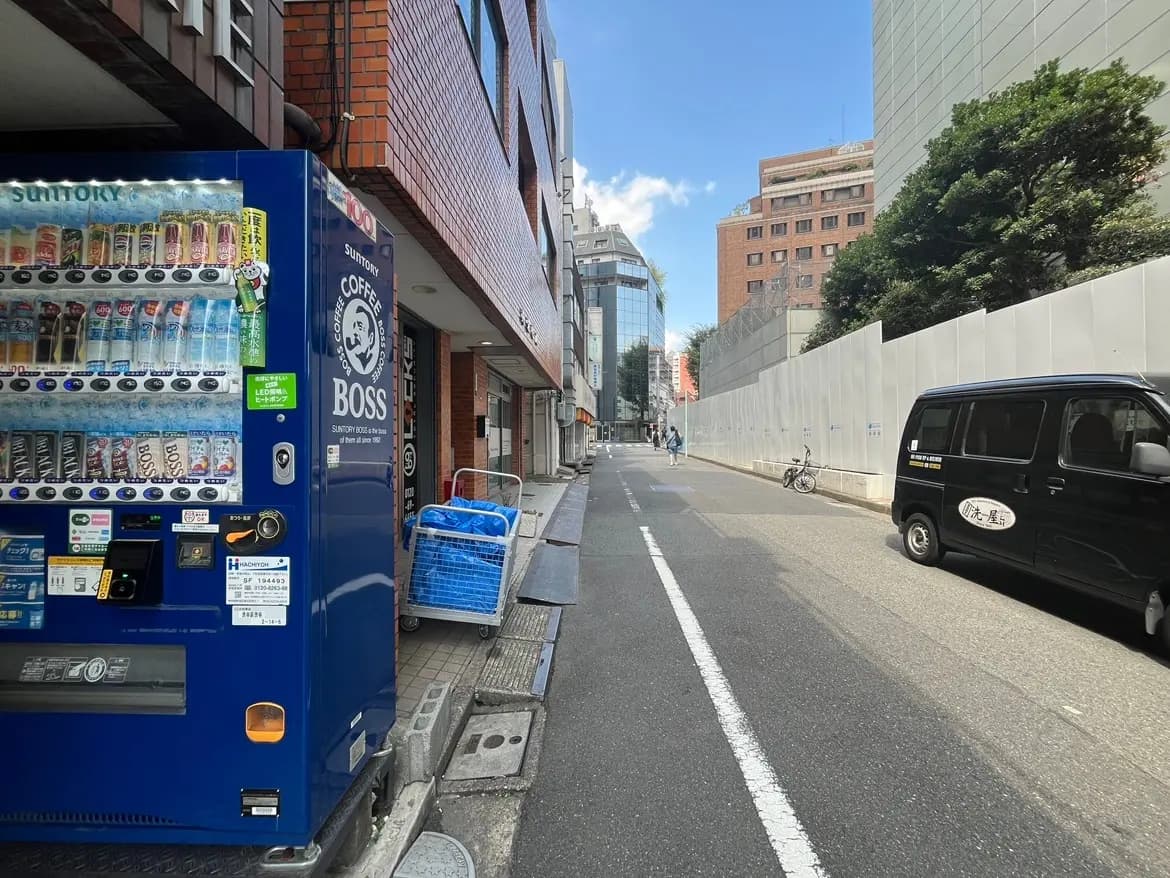 - サントリーの自動販売機、晴れた日を迎える東京の街角、建物が並ぶ通りと黒い車が走る風景