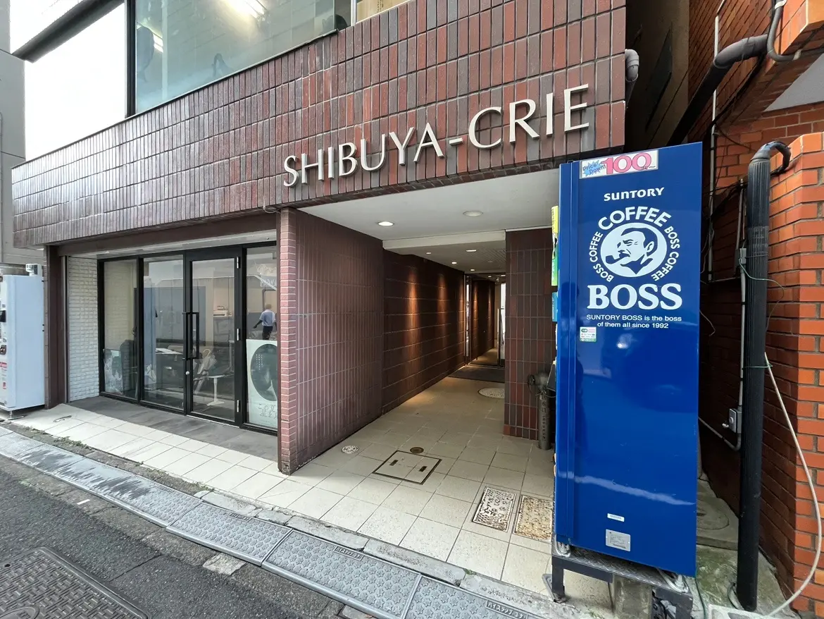 - 渋谷クリエのエントランス、ブリックタイルの外壁とガラスのドア、サントリーの自動販売機が置かれた街角の風景