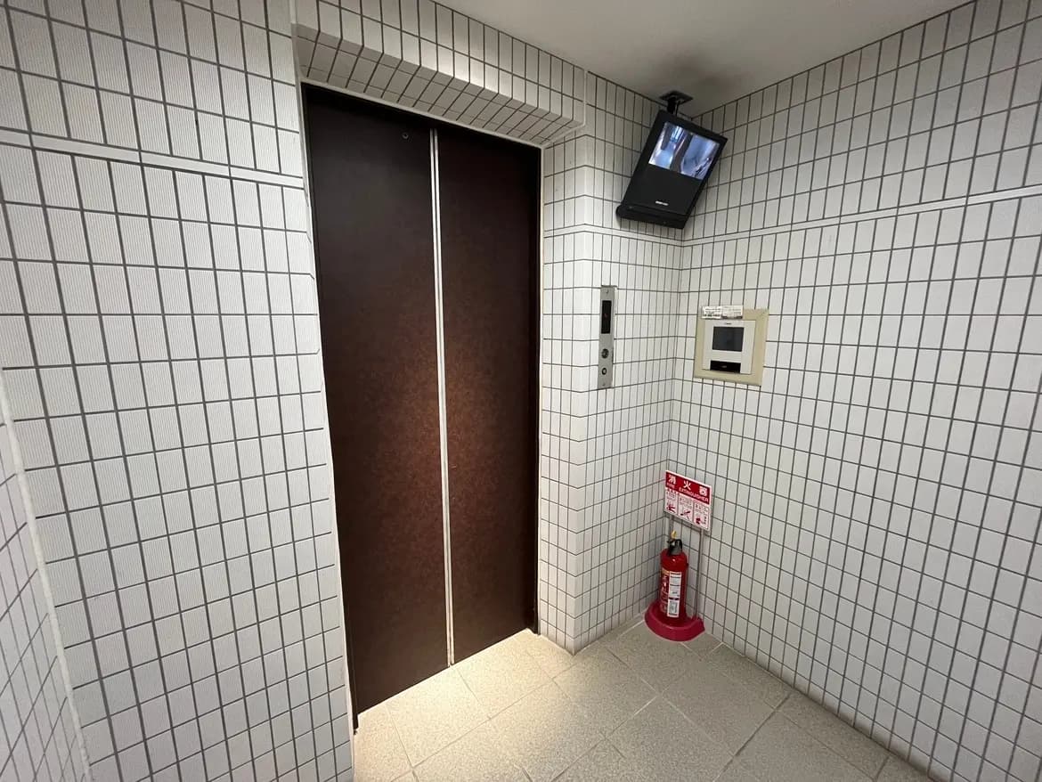 - 照明が明るいエレベーター前の待機スペース、タイル張りの壁に設置されたモニターと火災用消火器のある安全対策の環境