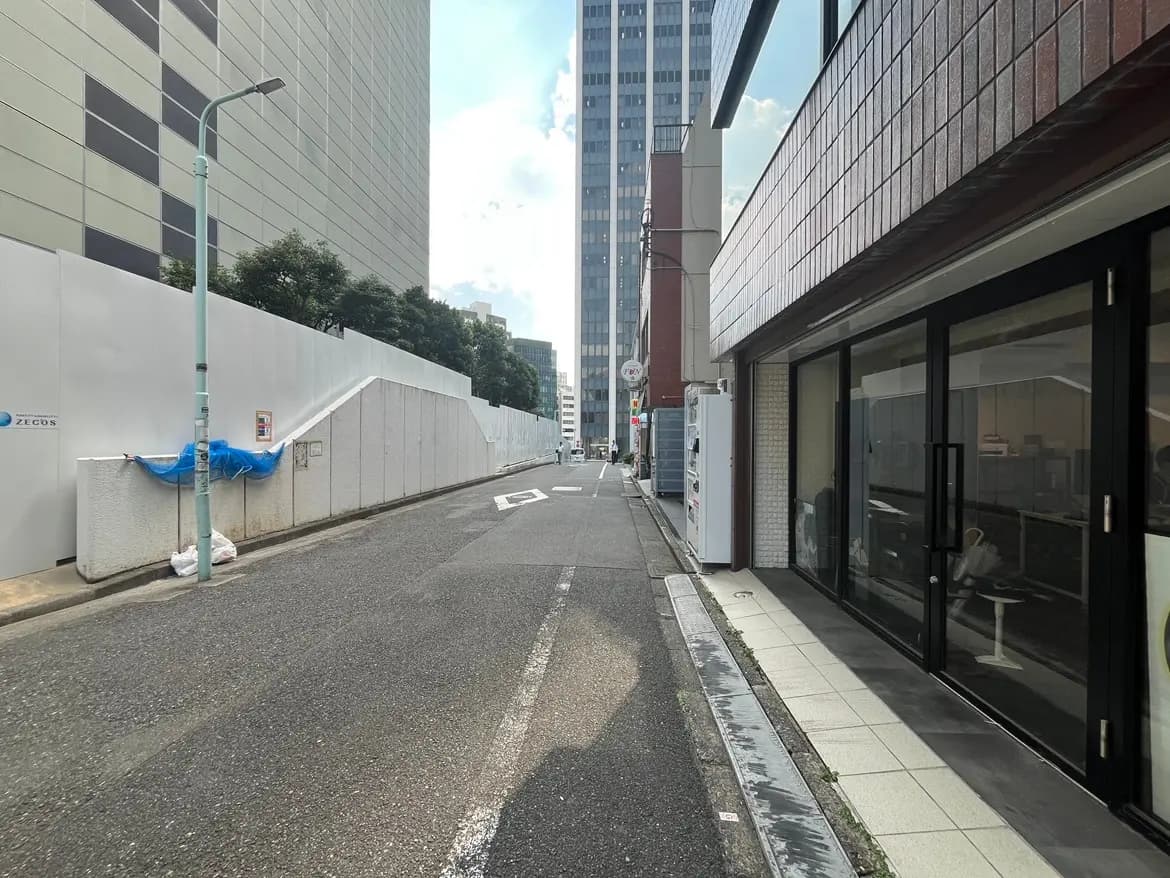 - 都市部の静かな通り、白い仮囲いとビルが並ぶ景色、青いシートと街灯が見える風景