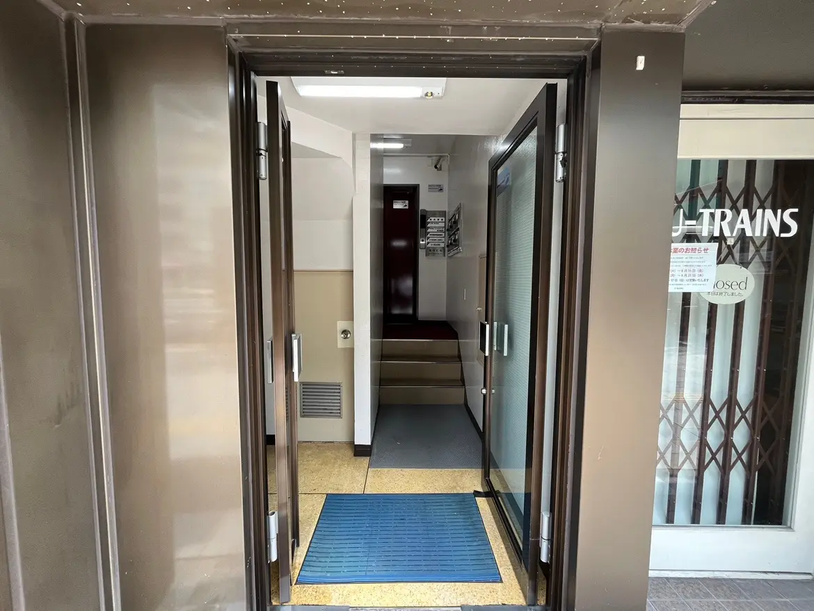 - 鋼鉄のドアが開いているエントランス、青いマットと階段が見える明るい受付エリア、隣に店舗の入り口がある景観