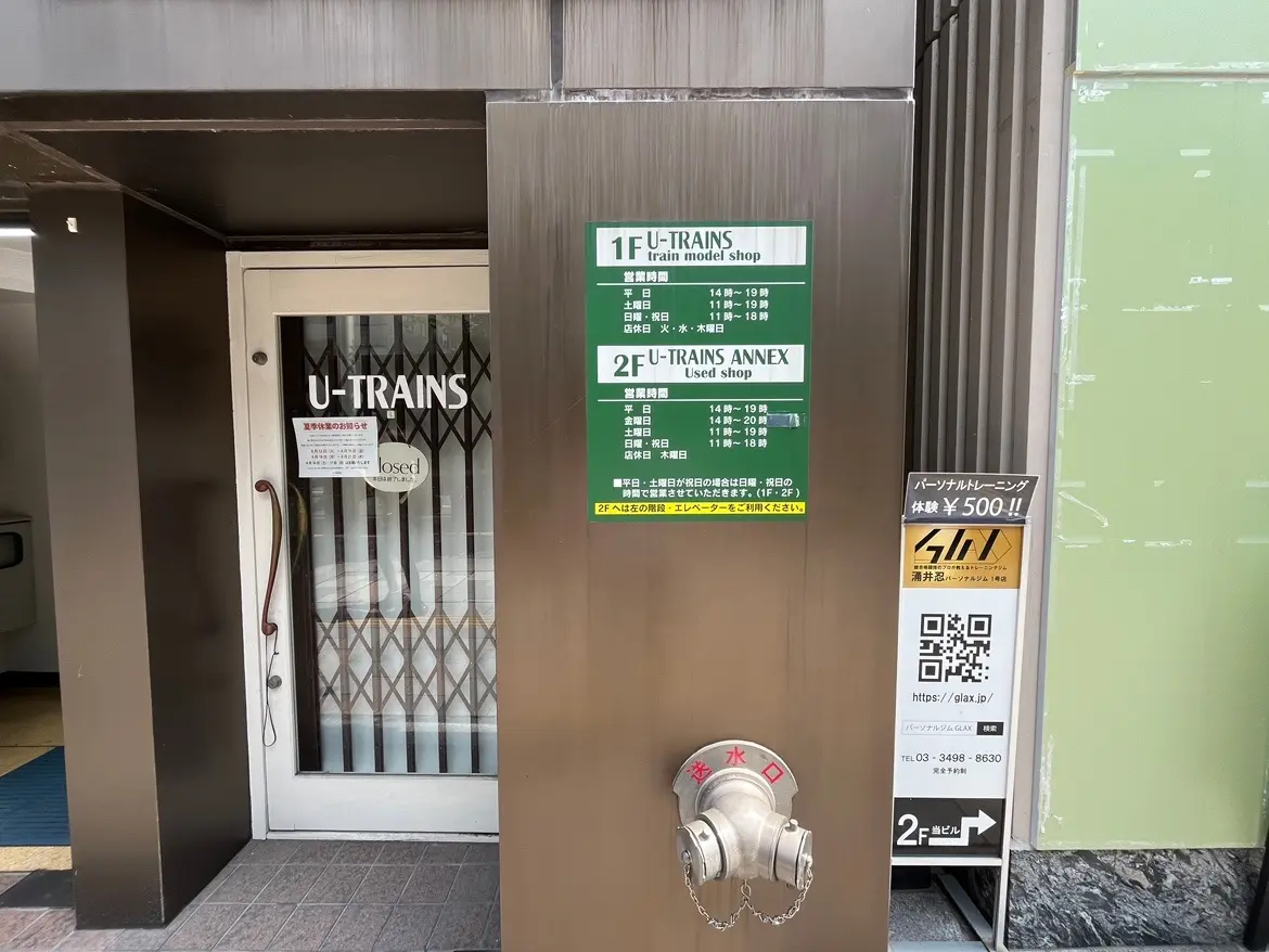 - 鉄道模型専門店「U-TRAINS」の入り口、営業時間案内が掲示された金属板と消防用水栓のある外観