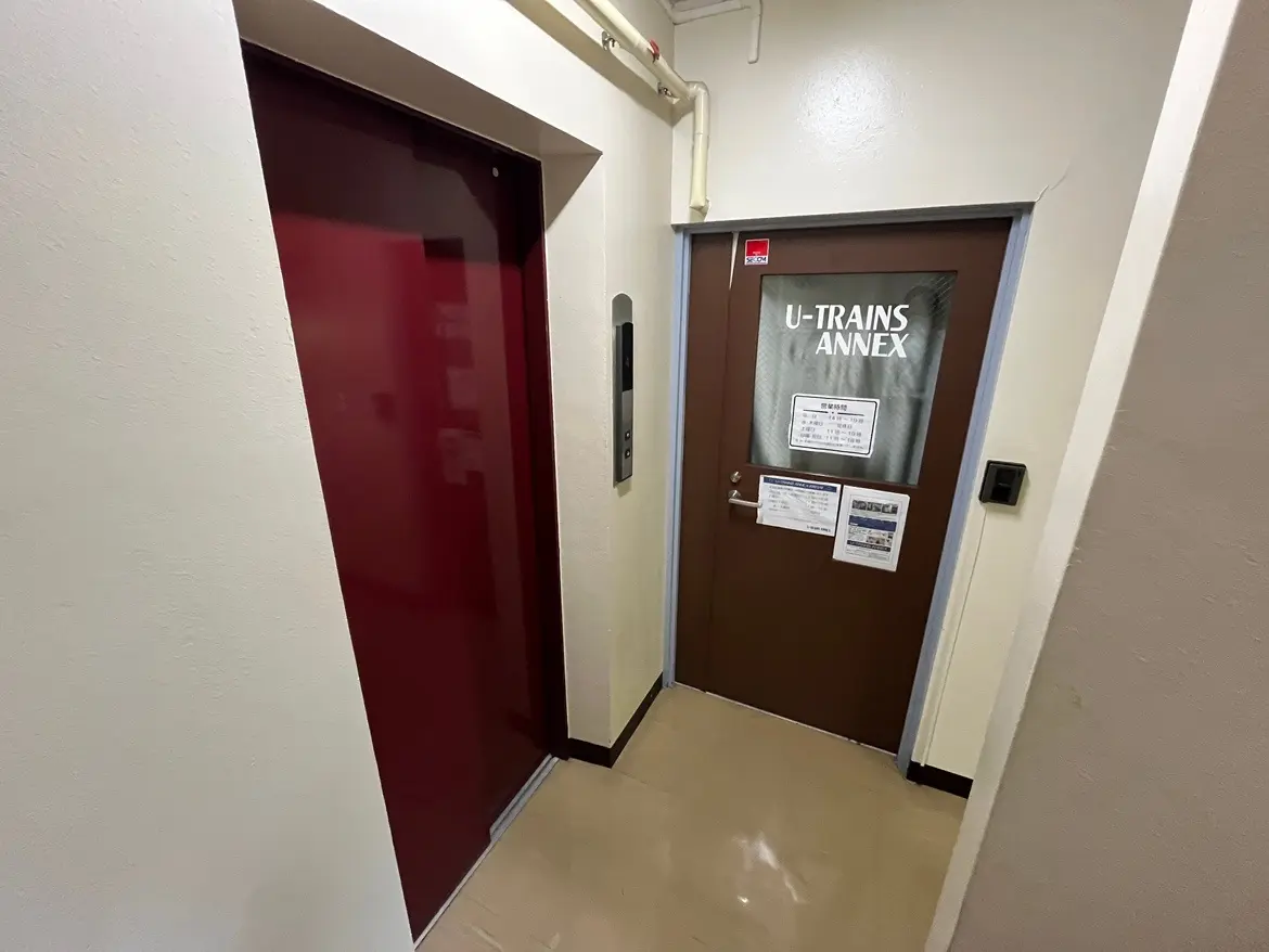 - エレベーターと「U-TRAINS ANNEX」と記されたドアがある廊下の風景、明るい壁とシンプルな内装が特色の空間