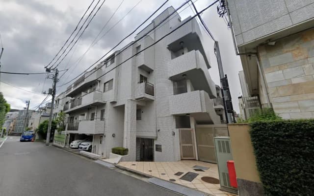 - 白い外壁のモダンなアパートメント、2階建てのバルコニーとエントランス、周囲に緑が広がる静かな住宅街の風景