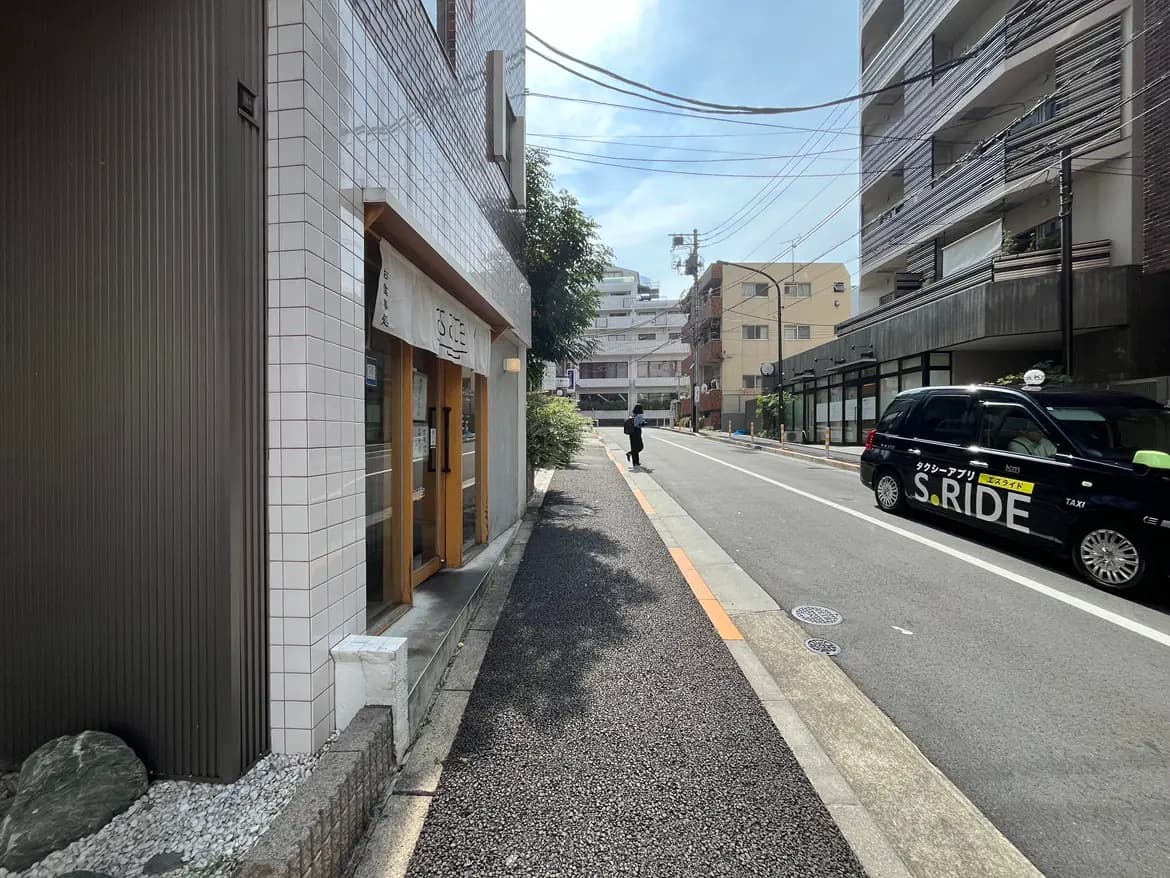 - 日差しが差し込む道沿いの風景、タクシーが停車し、並木道と周囲の建物が見える都市の一コマ