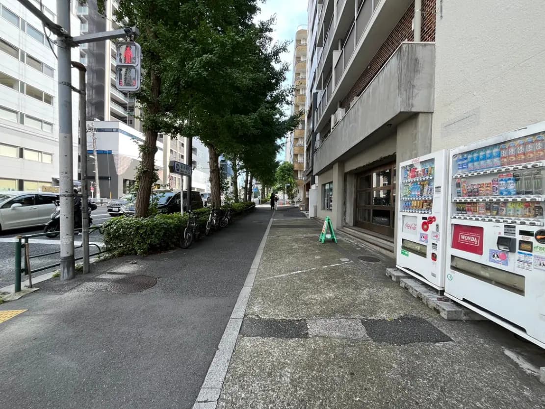 - 都市の歩道、緑の木々と自動販売機が並ぶ風景、車両通行の様子が見える通りの一角