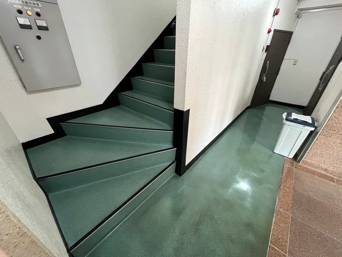 - 緑色の階段とフロアが特徴的な建物の内部、階段の横には郵便受けとゴミ箱があるエントランスホール