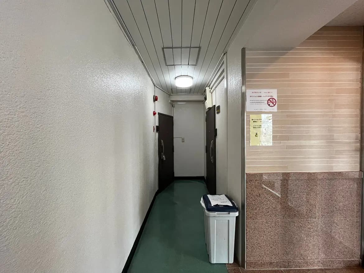 - 清潔感のある長い廊下、白い壁と緑のカーペット、両側にはドアが並ぶシンプルな内装、ゴミ箱と掲示板も視認可能