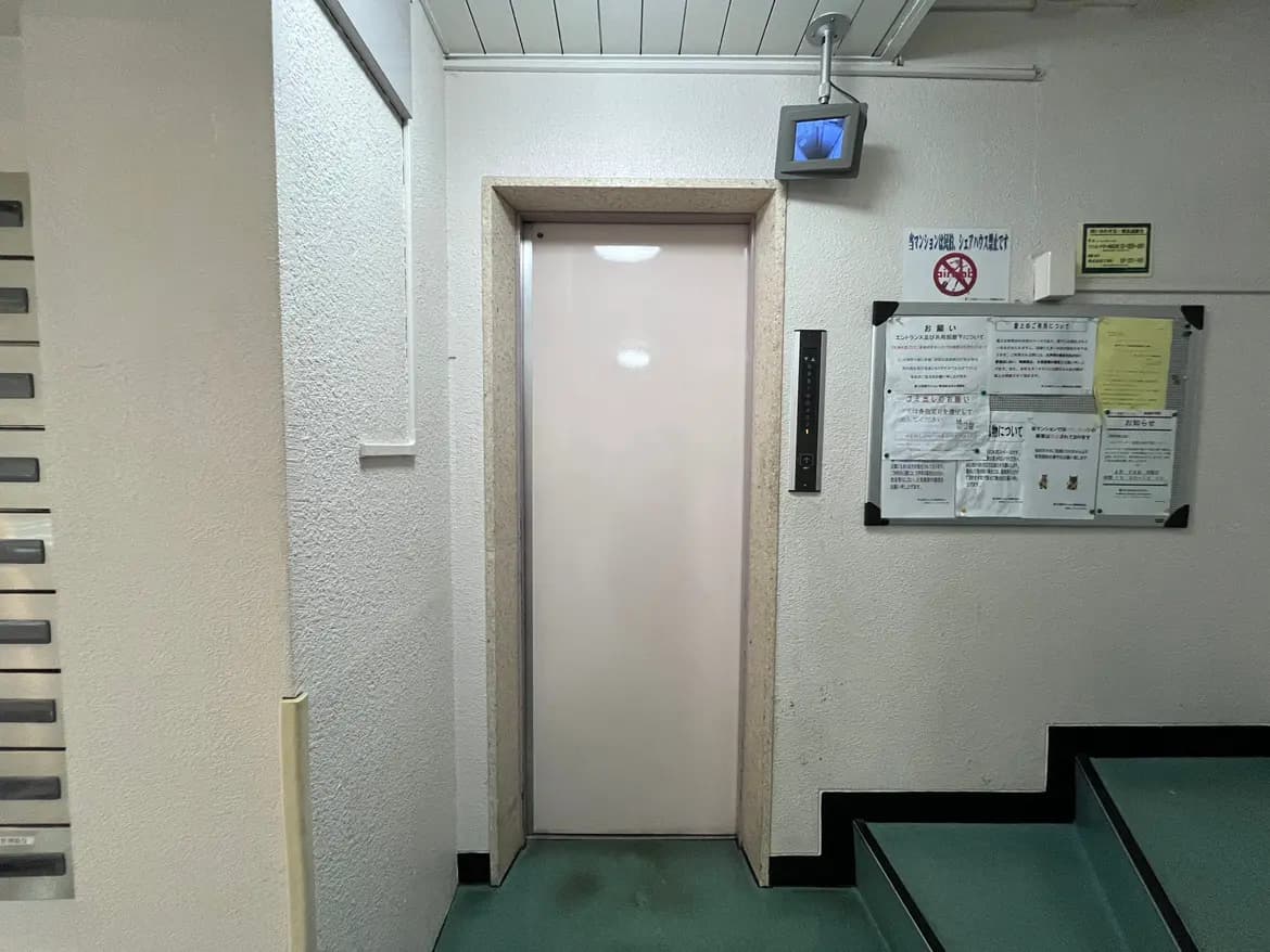 - エレベーターの入り口がある廊下の風景、壁に掲示板と監視カメラ、階段の手前にあるインターホン付きのエレベーターのドア