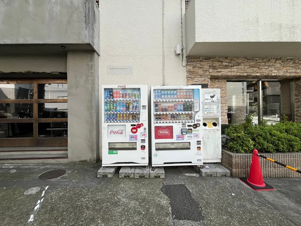 - コカ・コーラとWondaの自動販売機が並ぶ、雑多な雰囲気の街角、近くには緑の植栽とガラス張りの建物が見える景色