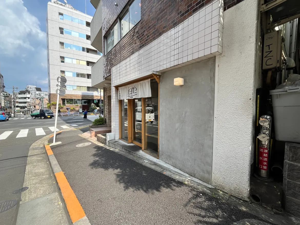 - 都市の交差点に位置する飲食店の外観、ガラス窓とシンプルな看板が特徴で、晴れた日に撮影された風景
