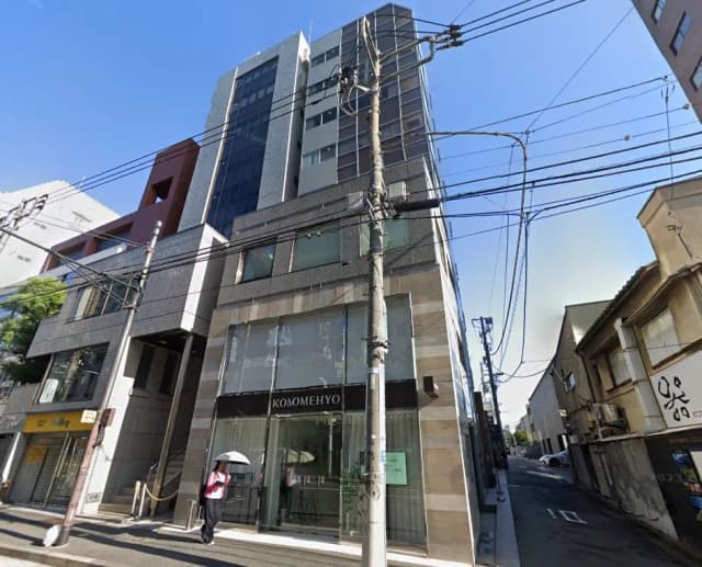 - 現代的なデザインのビル、ガラス張りのファサードと「COMOMEHYO」の看板、周囲の街並みが映る風景