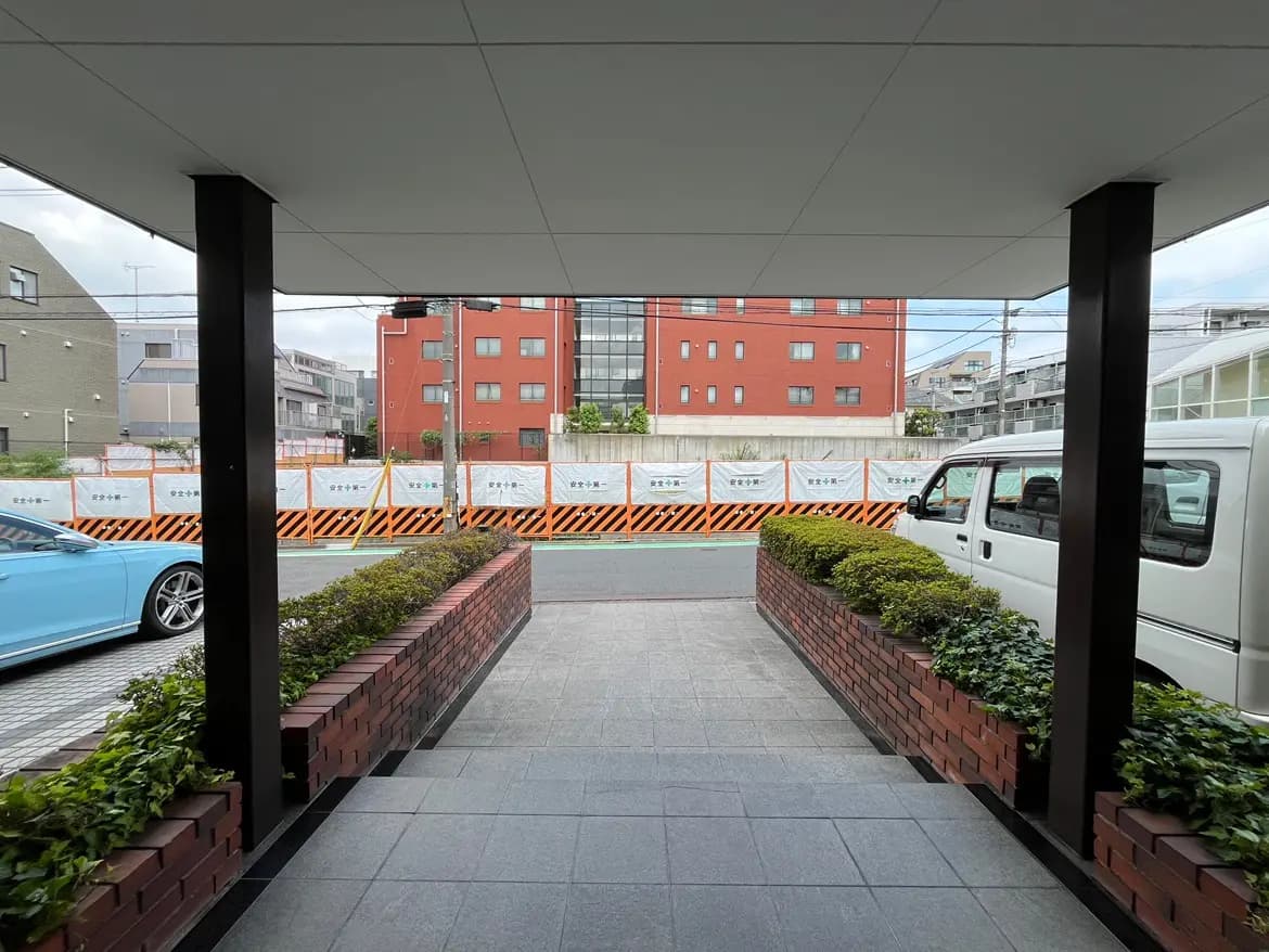 - 駐車場からのアプローチ、レンガ造りの花壇と緑が配置され、周囲には建物と工事のためのバリケードが見えるエントランスの風景