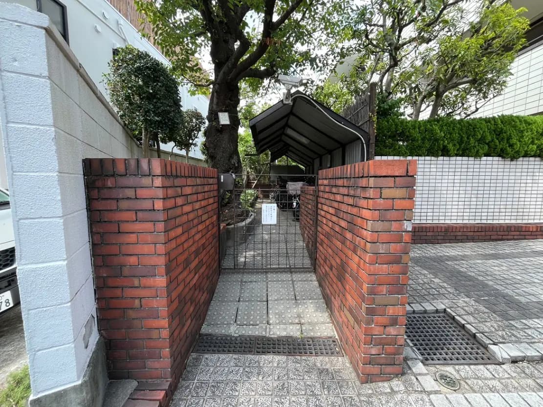 - レンガの壁で囲まれたエントランス、木々に囲まれたアプローチと屋根付きのスペースが見える玄関の景観