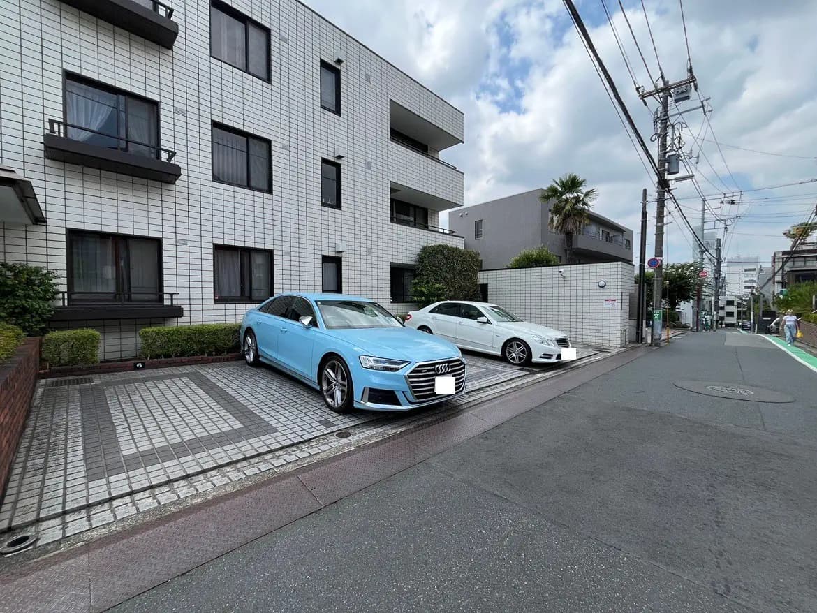 - 白い外壁のマンション前に駐車された青いアウディと白い車、周囲の緑と街並みが広がる風景