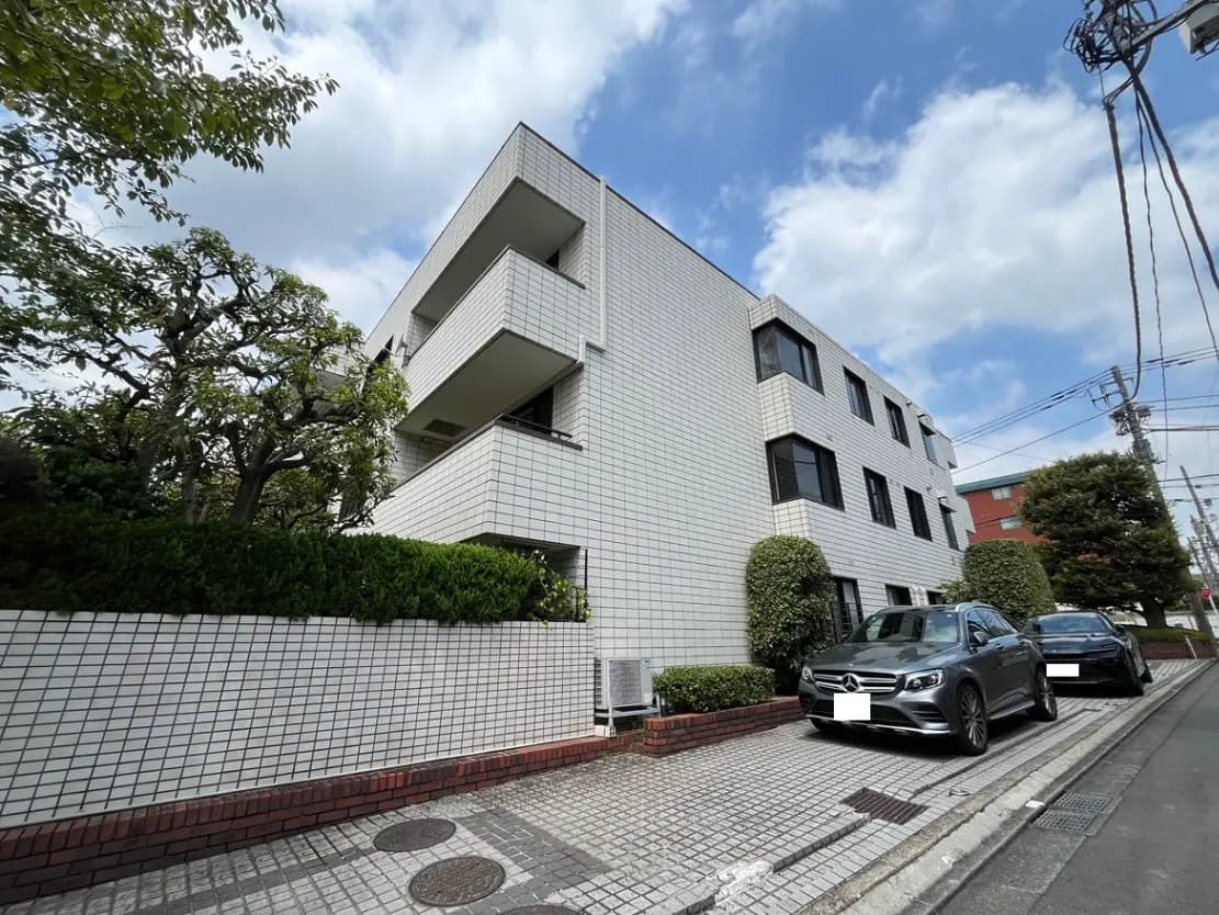 - 白いタイル張りのモダンなマンションと緑豊かな植栽、道沿いに駐車された車のある風景