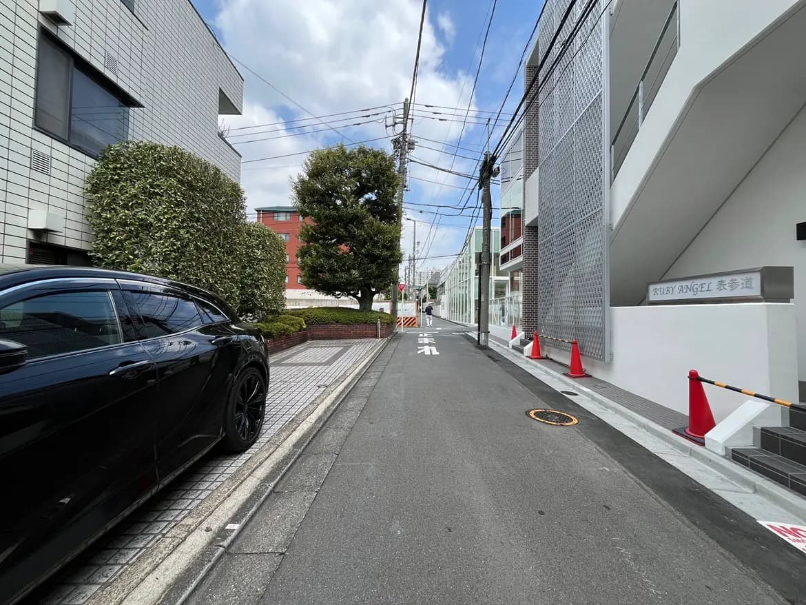- 緑豊かな街並みに面した狭い道路、黒い車と赤いコーンが配置された新しい建物の外観