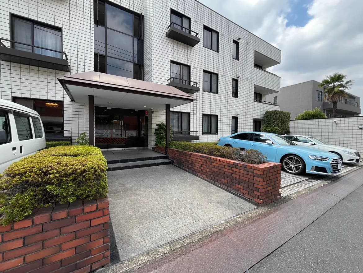 - モダンな外観の集合住宅、広々としたエントランス、整然と駐車された車と緑の植栽がある風景