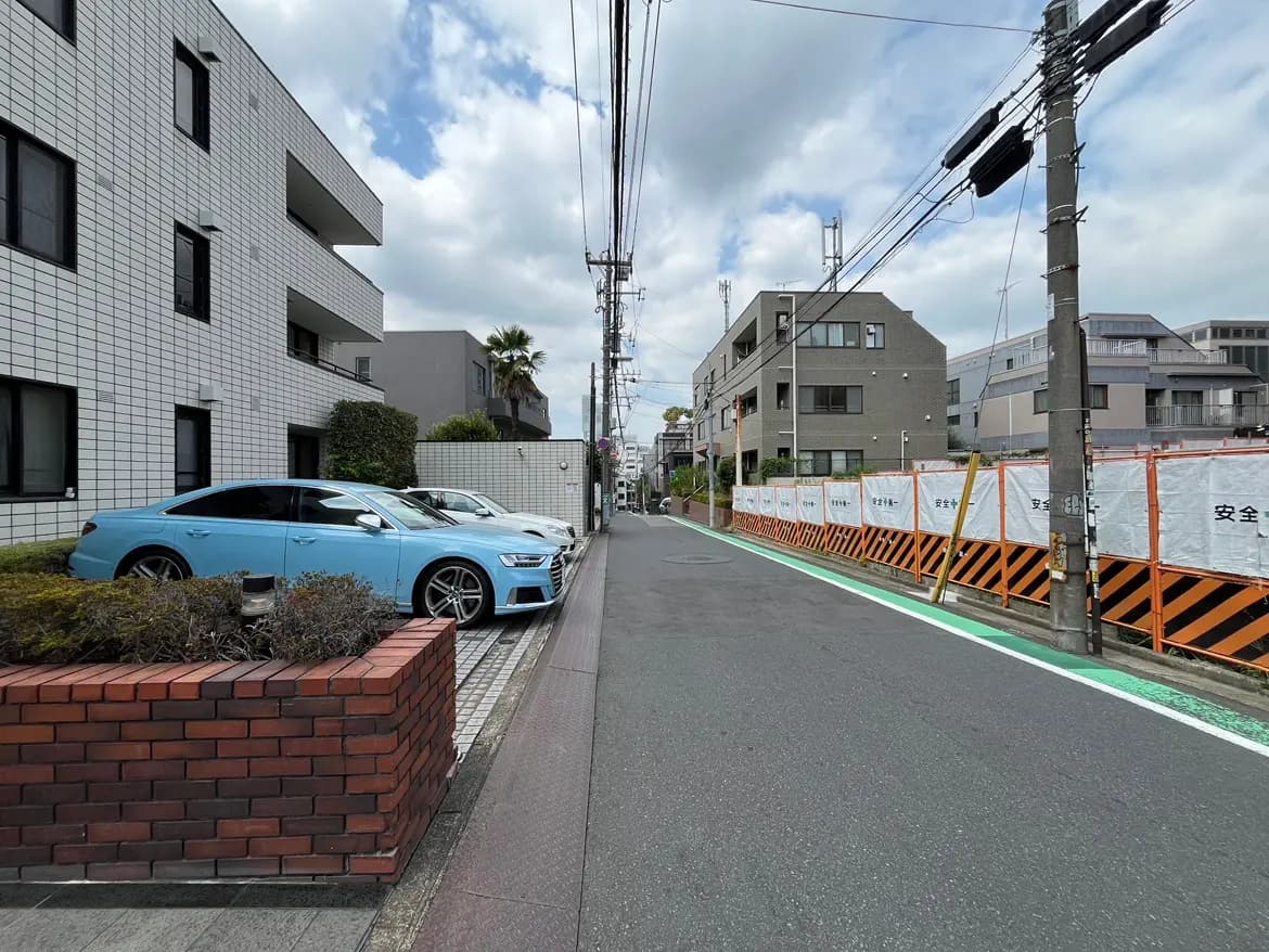 - 都会の住宅街、青い車が停まる歩道とブロック塀、舗装された道路や工事用のバリケードが見える風景