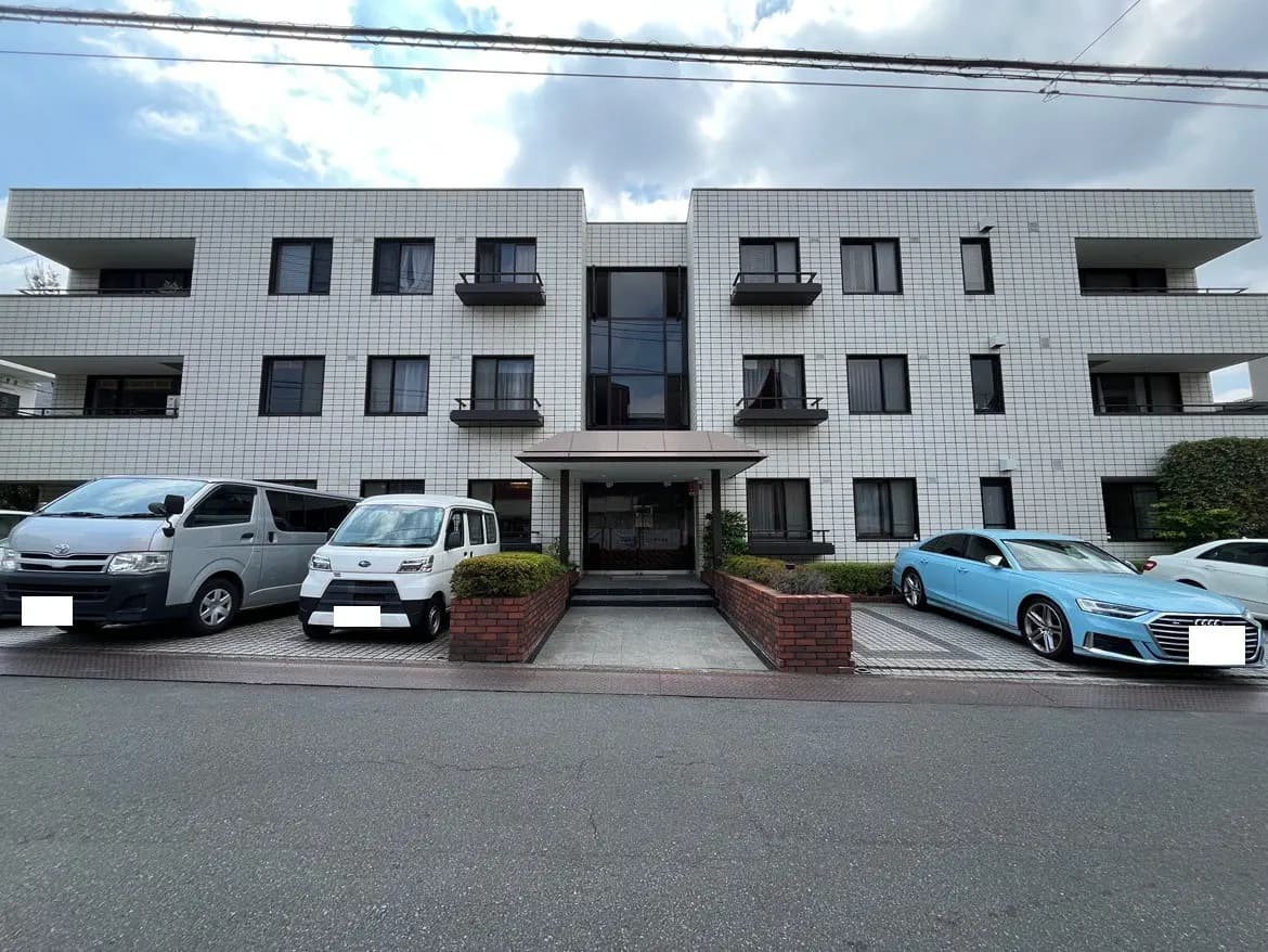 - シンプルでモダンな外観のマンション、目の前に駐車スペースがあり、青空の下に立つ建物の様子