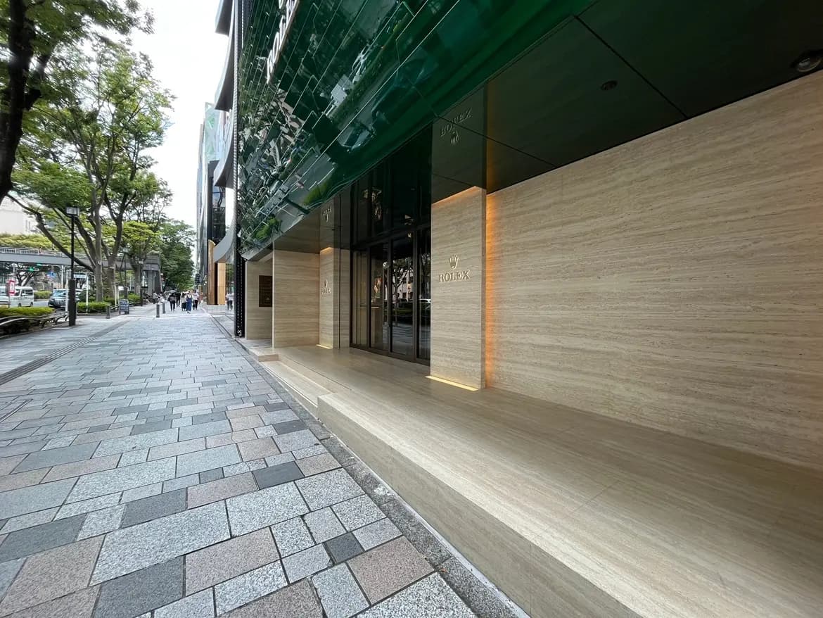 - 高級感のあるロレックスの店舗入り口、洗練された石材の外壁と緑のガラス屋根、通りには人々が行き交う風景