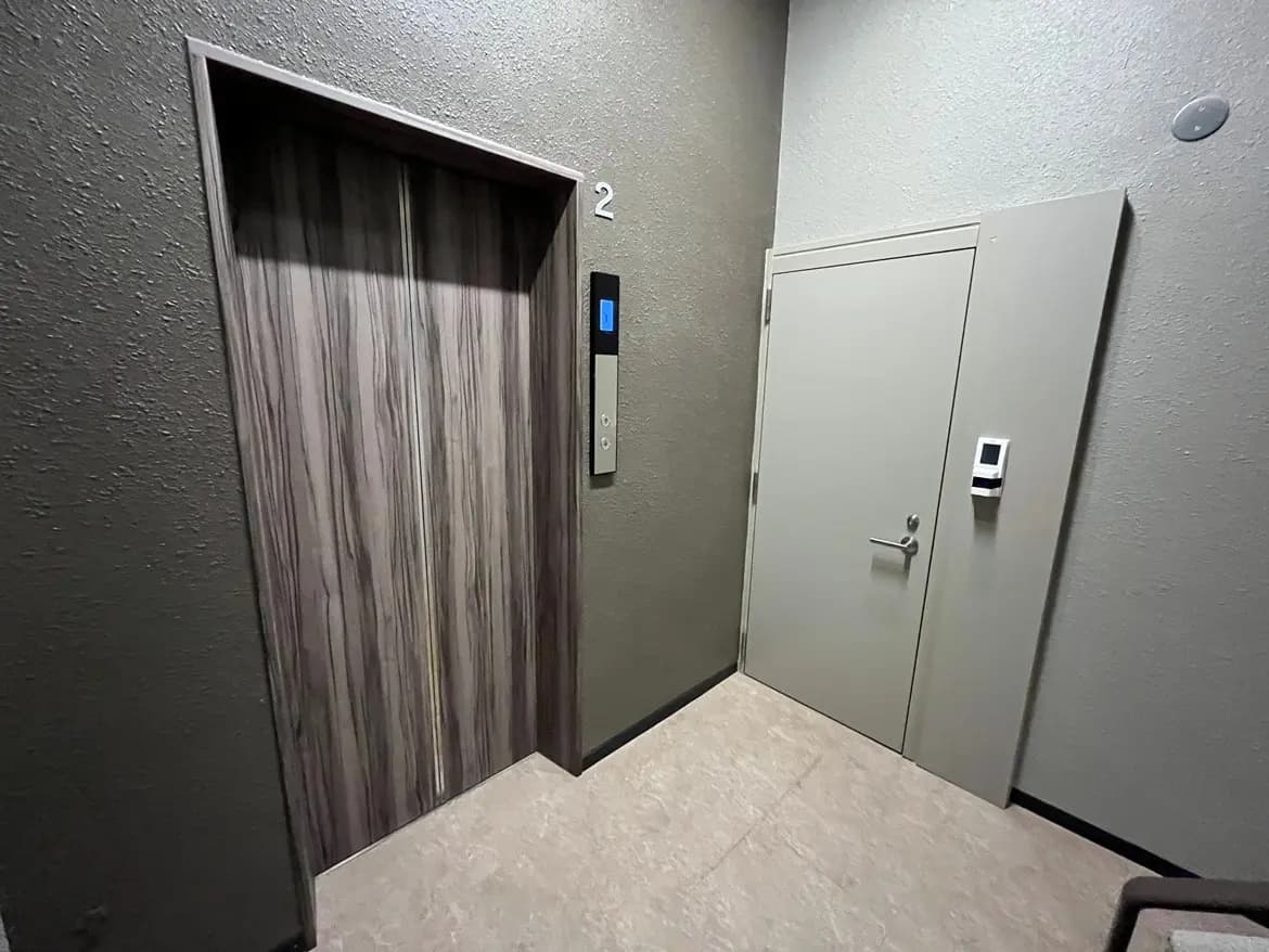 - コンパクトなエレベーターとドアがあるオフィスビルのエントランスホール、落ち着いたグレーの壁と明るいフローリングの flooring