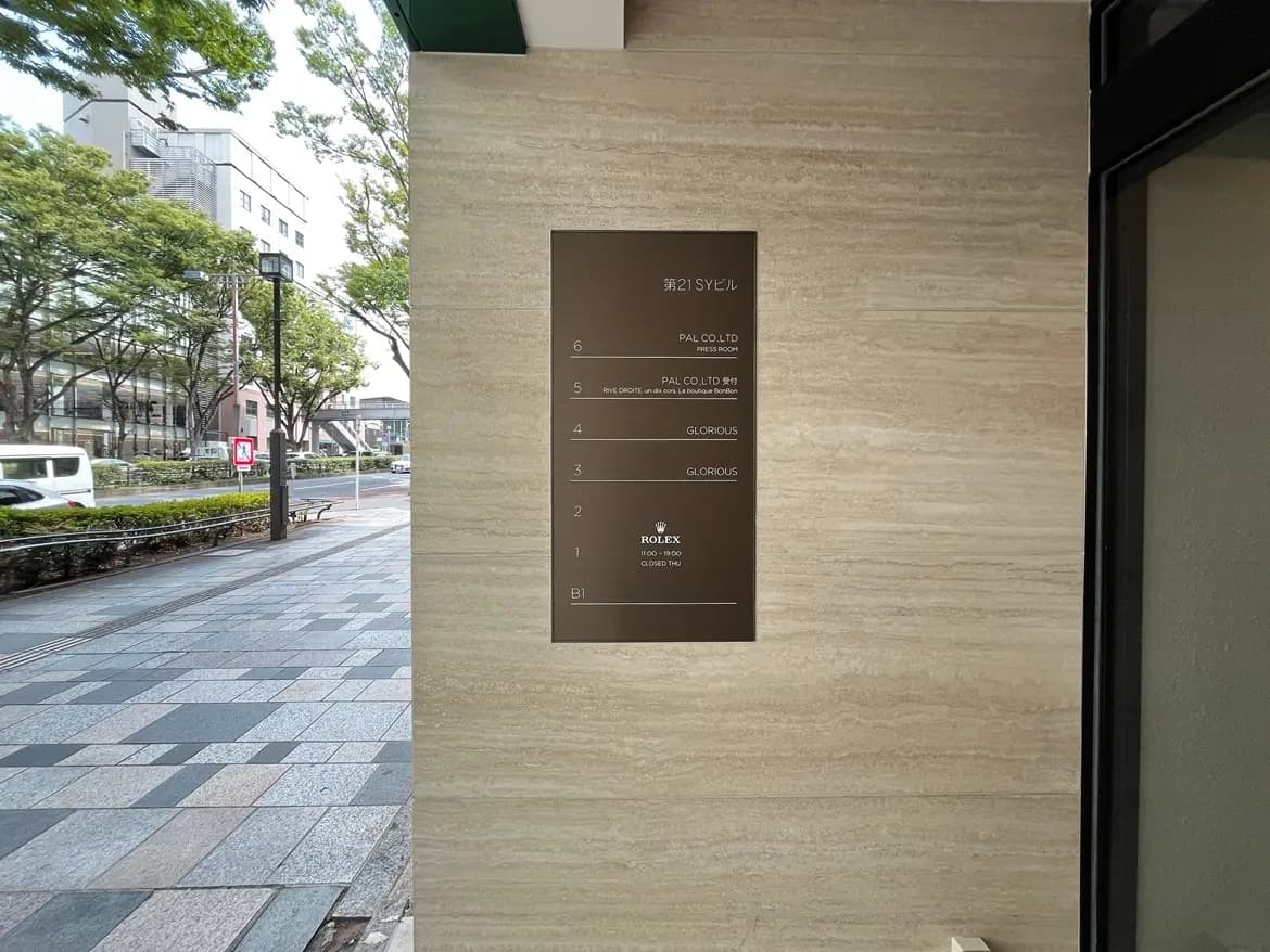 - 第21SYビルのエントランスサイン、テナント名が記載されたスタイリッシュな掲示板と周囲に広がる緑豊かな街並み