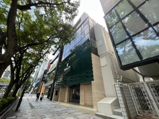 - 大きな窓と緑色の外装が特徴のロレックスの店舗、周囲には街路樹が並ぶ賑やかな通りの風景