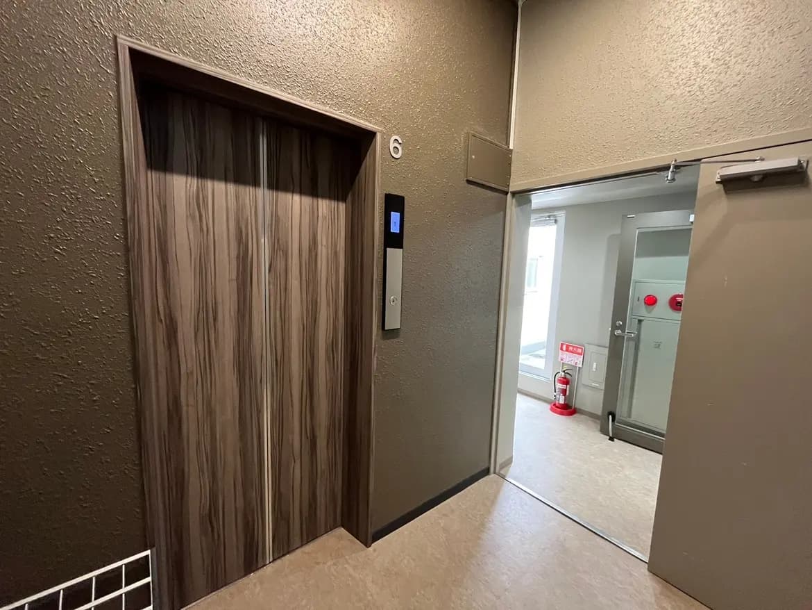 エレベーターのドアがある建物のエントランス、壁は暗い色合いで落ち着いた雰囲気、隣には窓と消火器が設置された出入口がある廊下のインテリア