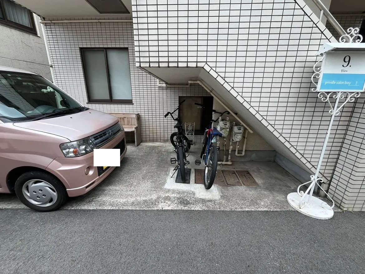 - 駐車スペースに停まったピンクの軽自動車と、自転車が停められた建物の入口、タイル貼りの壁とベンチがある風景