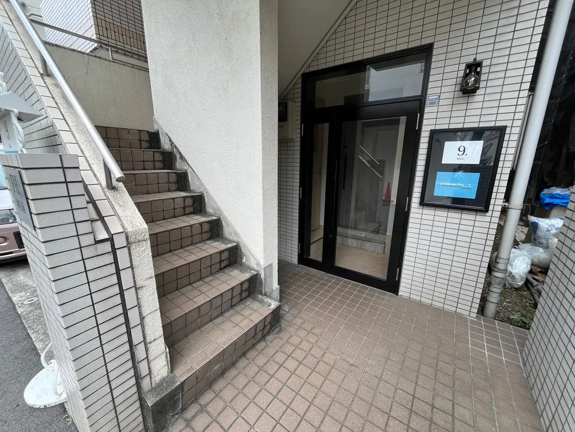 - 複数の階段が見えるマンションのエントランス、白い壁とタイル張りの床、ガラスの自動ドアが特徴の外観