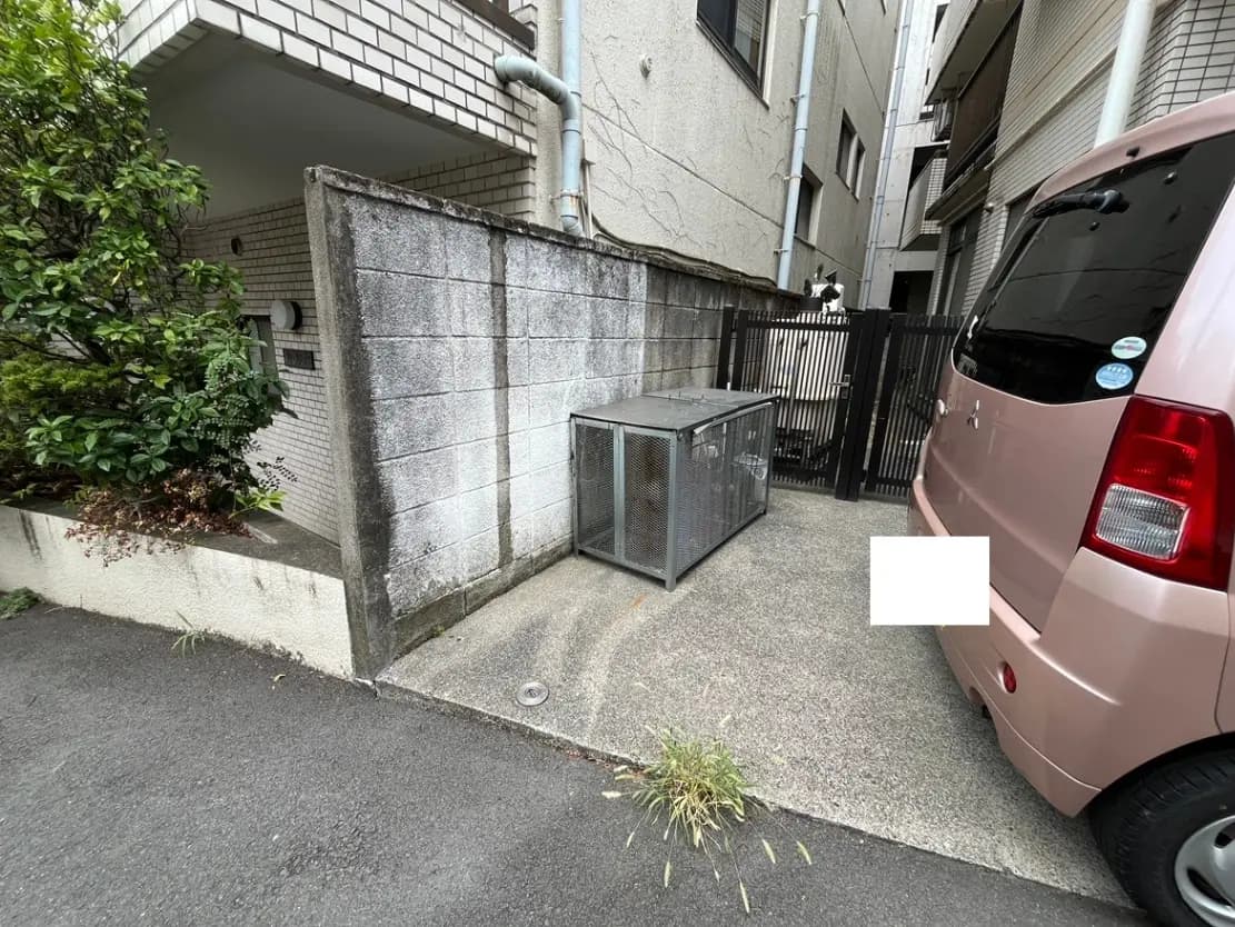 - 住宅の横に設置された金属製の収納ボックス、周囲に緑の植栽と舗装された道が見える駐車スペースの風景