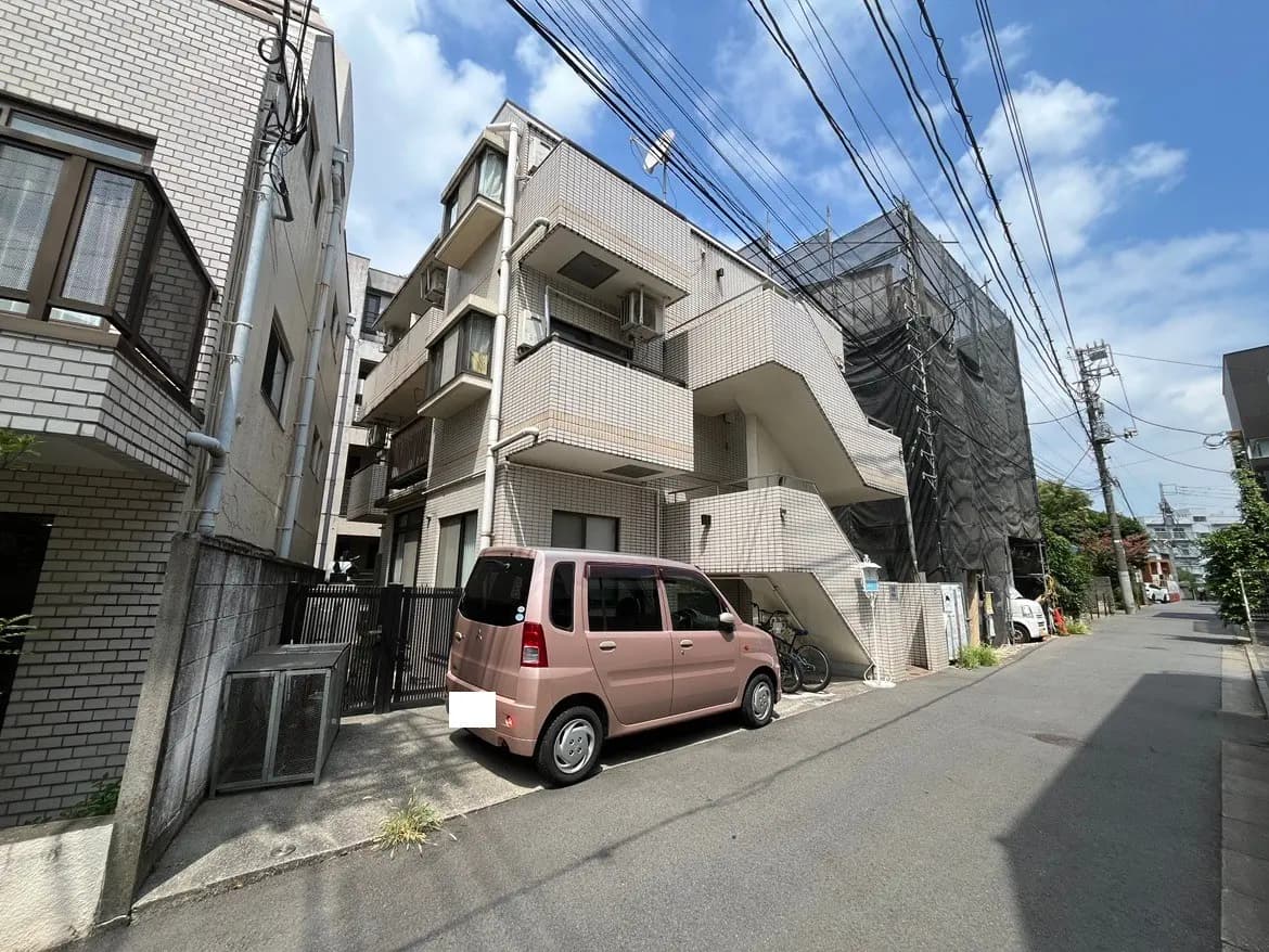 - 都市型のアパート外観、明るいレンガ調のタイル、駐車場に停まるピンクのコンパクトカー、周囲に住宅や電柱のある静かな通りの風景