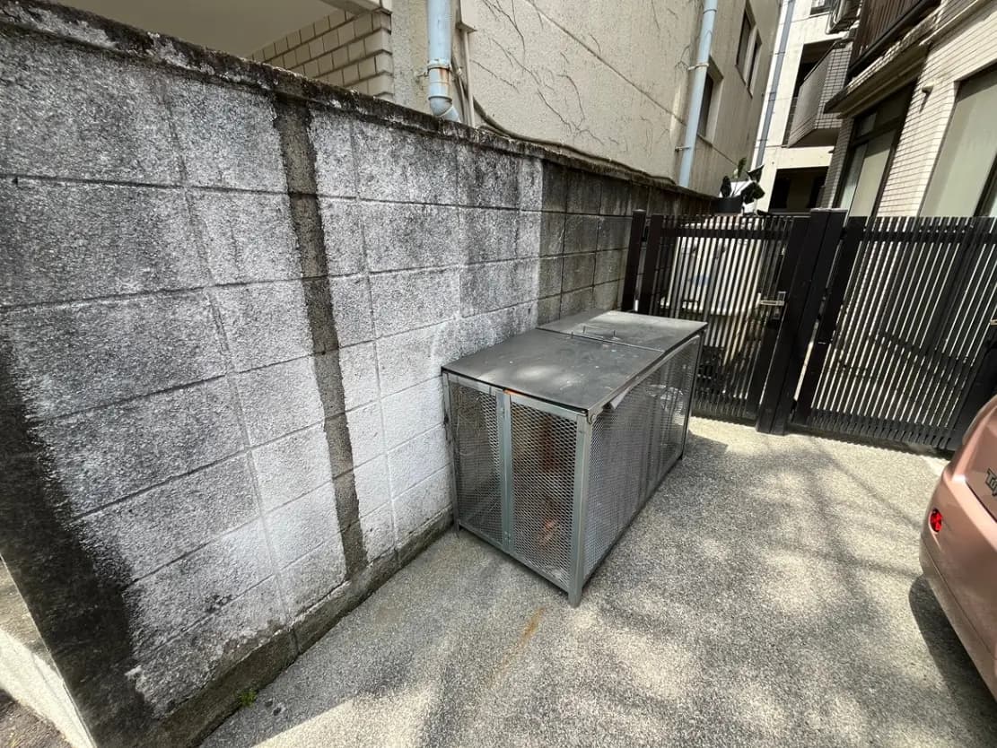 - グレーのブロック塀沿いに置かれた金属製のゴミ箱、周囲はコンクリートで舗装されている住宅街の風景