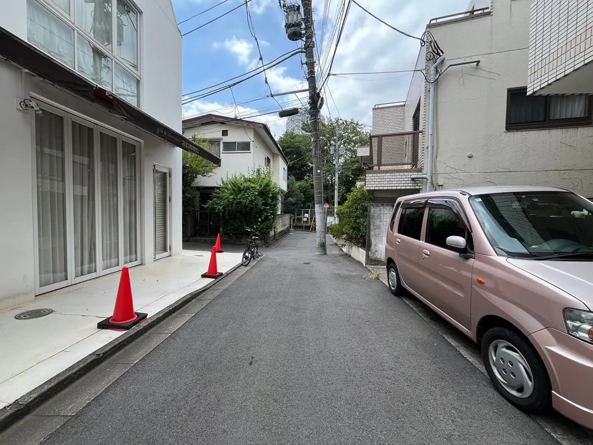 - 住宅街の静かな路地、白い外壁の建物と赤いコーン、ピンクの車と自転車が停まっている風景