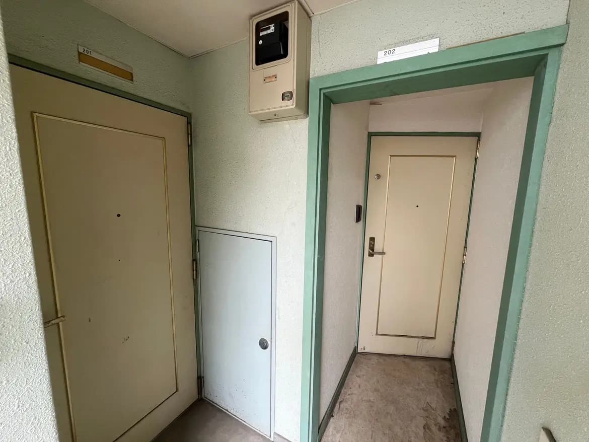 - 二つの扉が並ぶアパートの廊下、壁は淡い緑色で、上部には部屋番号のプレートが掲示されているシンプルな内装