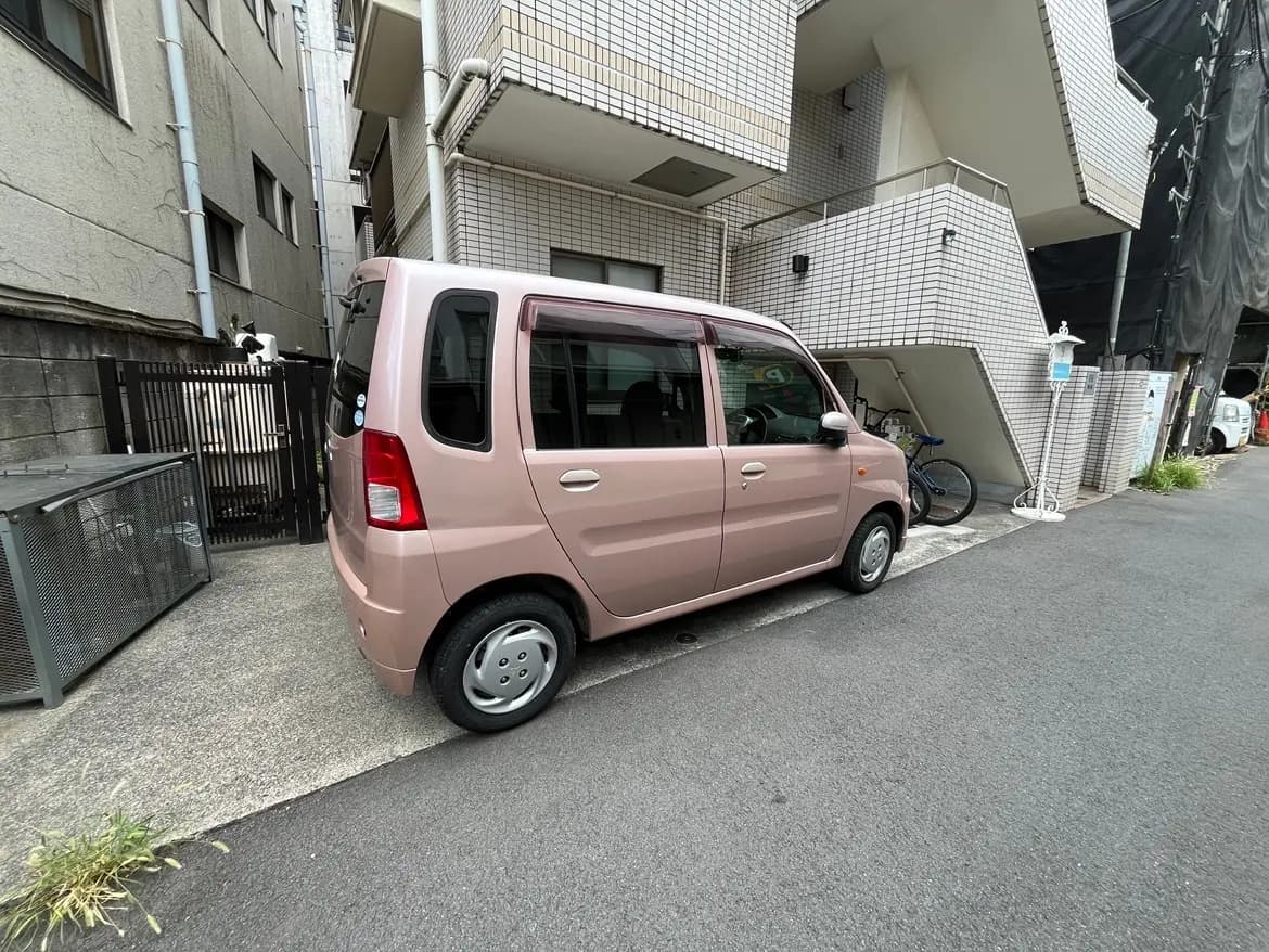- ピンクの軽自動車が都市の住宅街に駐車されている様子、周囲にはマンションと自転車が見える通りの風景