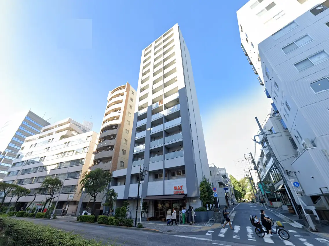 - 都会的な風景の中に立つ高層マンション、隣接する建物や緑地、青空が広がる通りの景観
