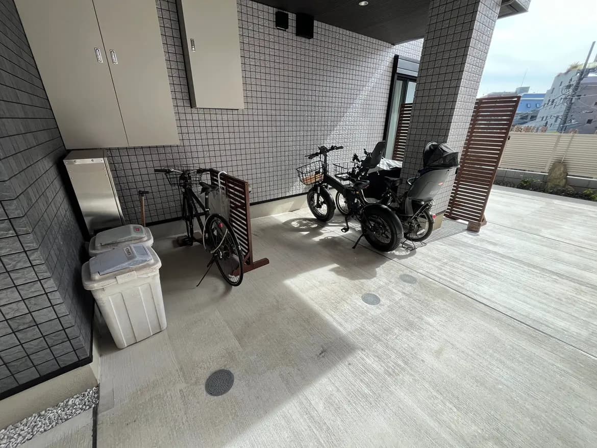 - 住宅の外にある自転車置き場、二台の自転車と赤ちゃん用のカーゴバイク、ゴミ箱と収納が配置されたシンプルなスペース