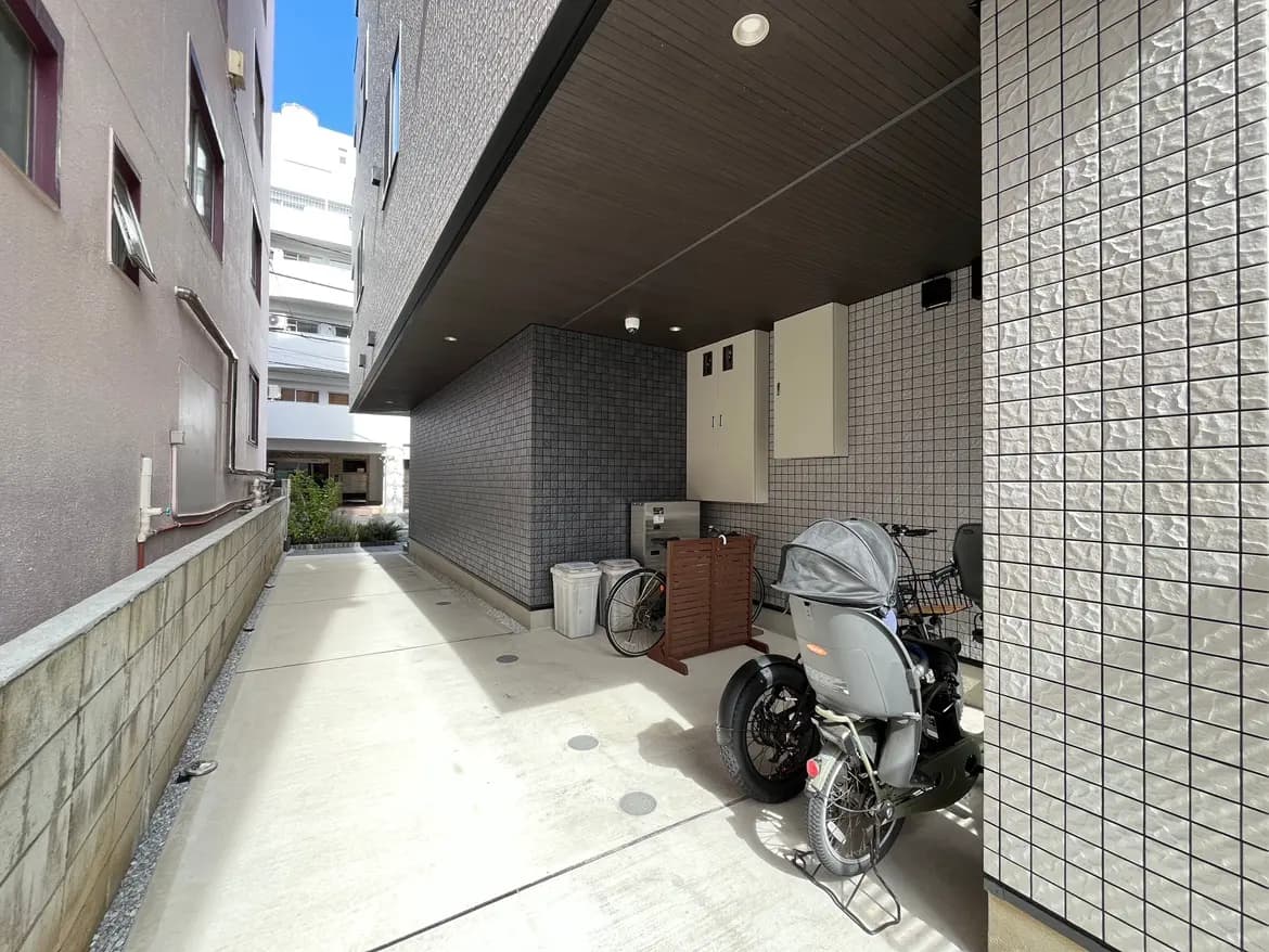 - 洗練されたデザインの屋外スペース、自転車とゴミ箱が置かれた清潔感のある通路、隣接する建物の壁が写る景観