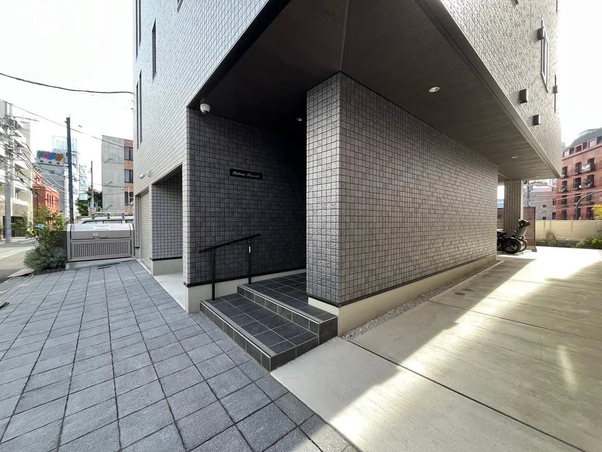 - 現代的なデザインの建物エントランス、タイル張りの壁と階段、周囲の街並みが見える風景