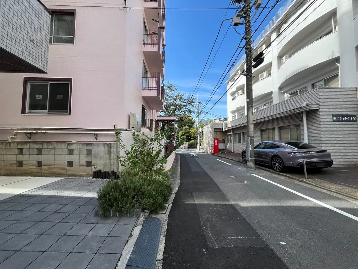 - ピンクの外壁のアパートと白い建物が並ぶ街並み、青空と電柱が見える静かな通りの風景