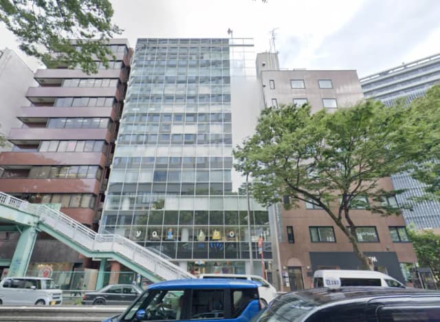 - ガラス張りの現代的なオフィスビル、隣接する低層ビルと緑の木々がある風景、道路沿いの駐車車両が見える