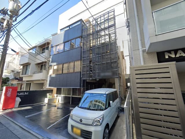- ビルの外観、足場が組まれた建物と駐車スペースに停まる白い軽自動車、青空が広がる都市の風景