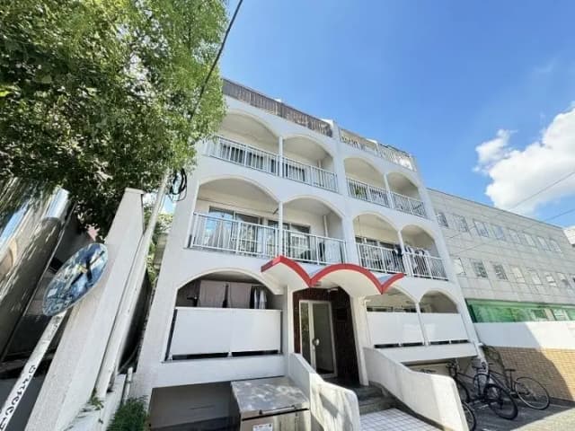 - 明るい青空の下で見える白い外壁の住宅ビル、アーチ型のバルコニーと赤い屋根が特徴、周囲に植木がある閑静な環境