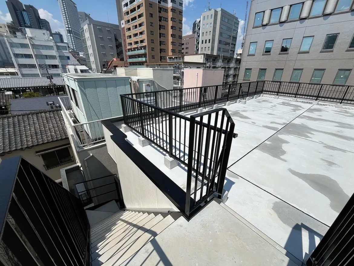 - 都市の屋上テラス、ステップでアクセス可能な白いコンクリートの床と黒い手すり、周囲の高層ビルが見える風景